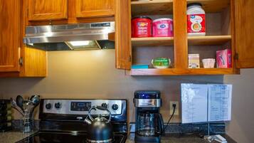 A kitchen with wooden cabinets, a stove, a kettle, a coffee maker, and various food items on shelves.
