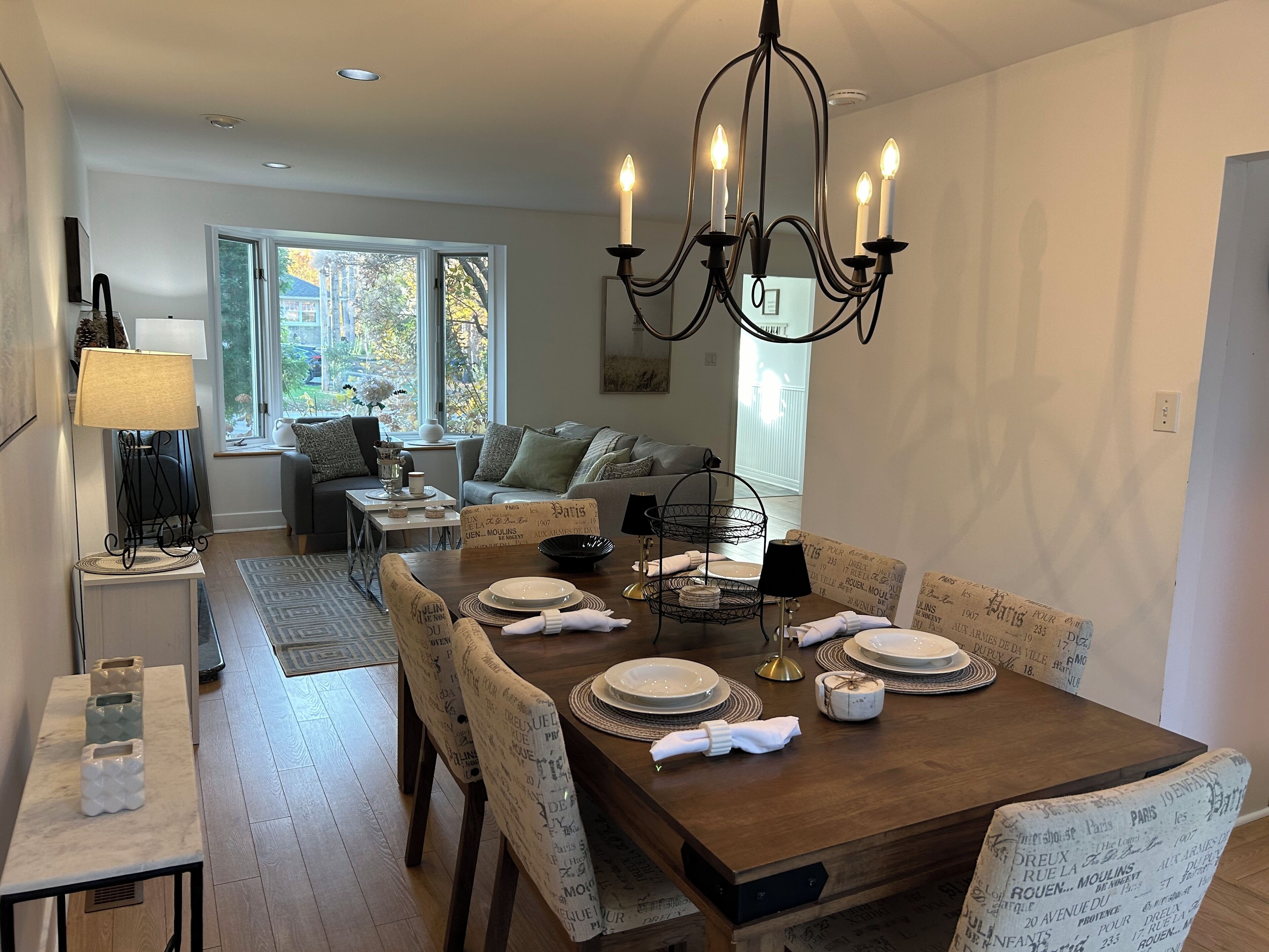 Un coin repas avec une table en bois dressée pour quatre personnes, entourée de chaises à motifs. Un lustre est suspendu au-dessus, et une fenêtre laisse entrer la lumière naturelle.