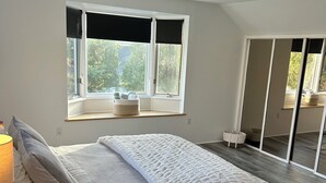 Et moderne soveværelse med en stor seng, et vindue med udsigt til træer, et sengebord med en lampe og trægulv.