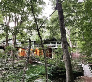 Eine Hütte, umgeben von dichtem Wald, mit einer Holzterrasse und einer Treppe, die zu einer zweiten Ebene führt.