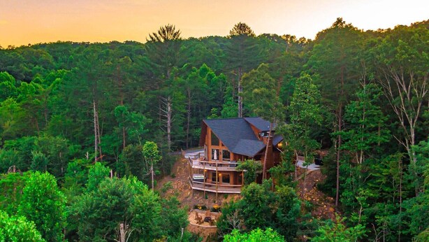 Una cabaña rodeada de bosque frondoso, con una estructura de madera destacada y un balcón con vistas a los árboles.