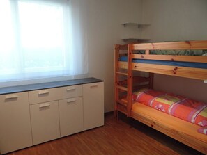 Una cama litera con estructura de madera y colcha colorida, un armario blanco con varios cajones y una ventana con persianas.