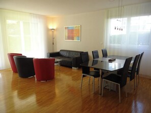 Ein modernes Wohnzimmer mit einer Couch, roten Sesseln, einem Esstisch mit Stühlen und einem Gemälde an der Wand.