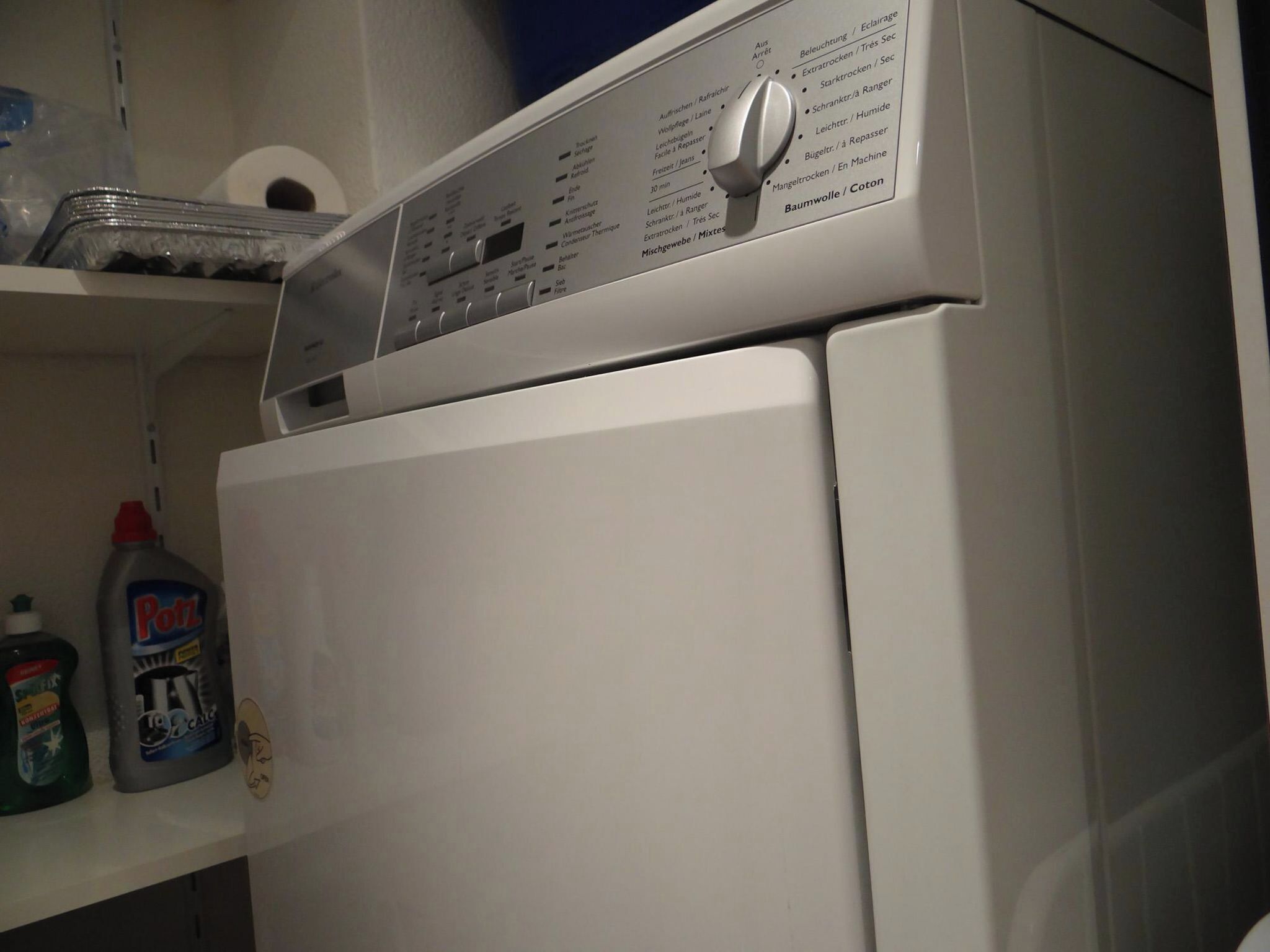 Une machine à laver blanche, dotée d’un panneau de commande et d’une étagère au-dessus, où sont rangés les produits de nettoyage.