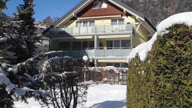 Un bâtiment à deux étages doté d’un balcon, entouré d’arbres couverts de neige et avec une chaîne de montagnes en arrière-plan.