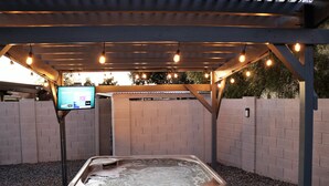 Un área de hidromasaje al aire libre con una pérgola de madera y luces colgantes, rodeada de muros de ladrillo y grava.