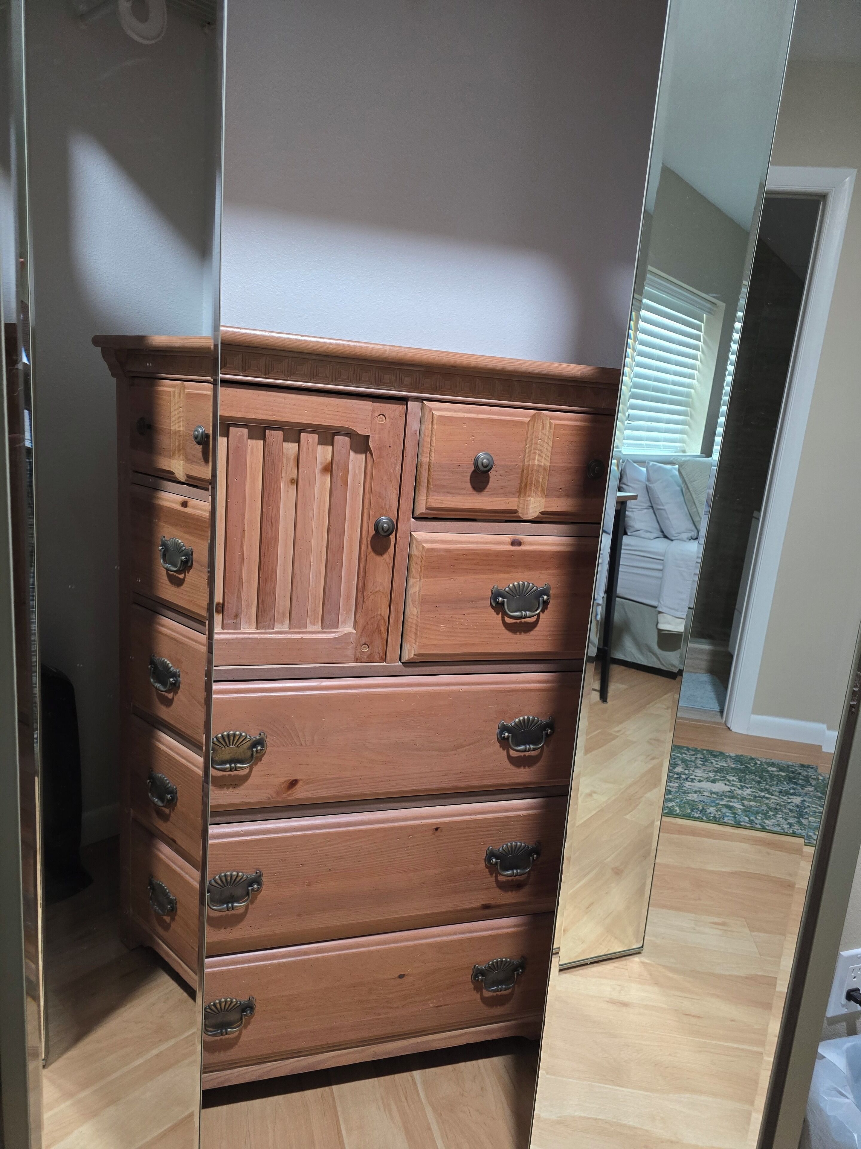 A wooden dresser with multiple drawers and knobs, positioned in a room with a wooden floor and a mirror reflecting part of the room.