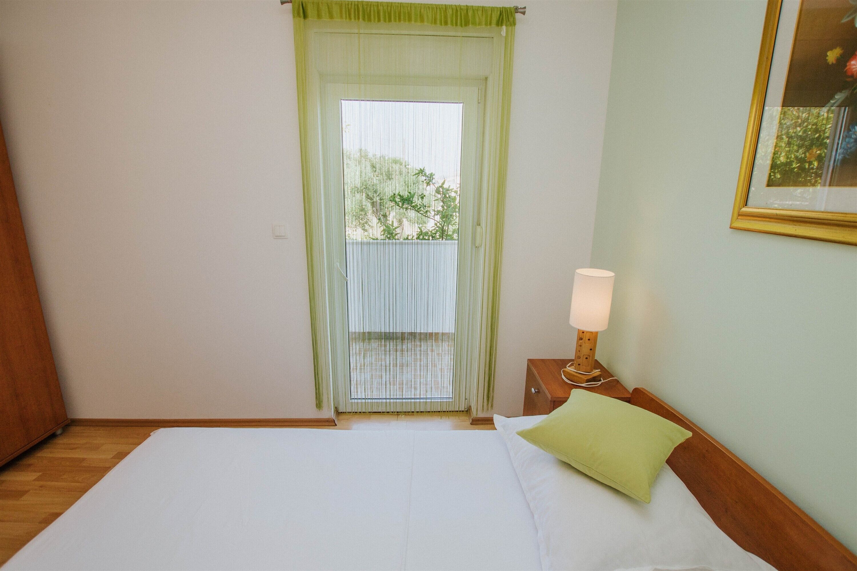 A bedroom with a bed, a nightstand with a lamp, a window with a view of greenery, and a framed picture on the wall.