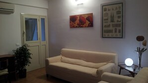 Een woonkamer met een beige bank, een potplant, een lijst met foto en een wandairconditioner.