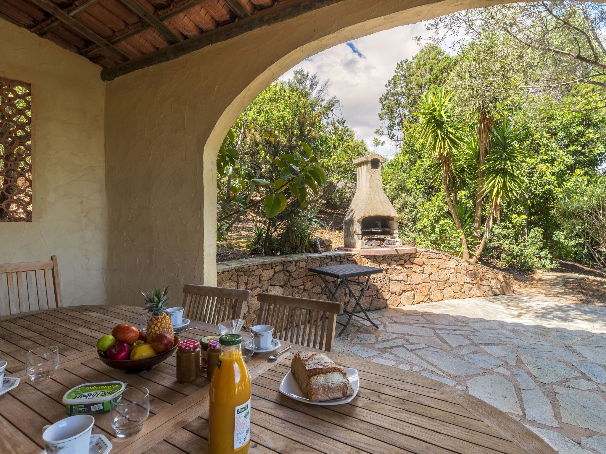 An outdoor dining area with a stone table, chairs, a fruit bowl, a bottle of juice, and a bread plate.