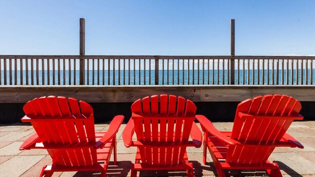 Three red Adirondack chairs facing a wooden deck with a railing overlooking a body of water. - Unwind at the Scallop Saturday cottage located at the Oceana Cottages (North Truro)