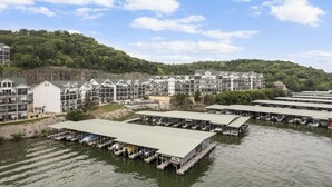 Un hotel frente al agua con varias embarcaciones amarradas, un edificio grande con techo plano y una colina boscosa al fondo.