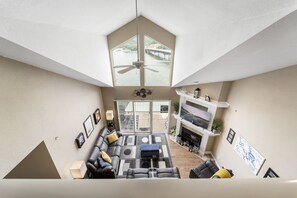 A spacious living area with a high ceiling, a ceiling fan, a large window with a view of a bridge, a fireplace, and a sectional sofa.