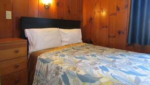 A bedroom with wooden walls, a bed with a floral bedspread, a nightstand, and a wall-mounted lamp.