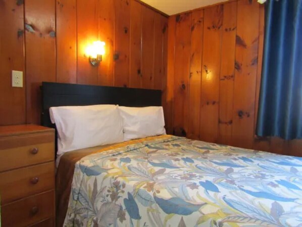 A bedroom with wooden walls, a bed with a floral bedspread, a nightstand, and a wall-mounted lamp.
