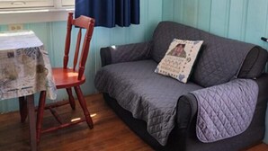 A room with a wooden floor, a blue wall, a grey sofa with a quilted cover, a wooden chair, and a table with a patterned tablecloth.