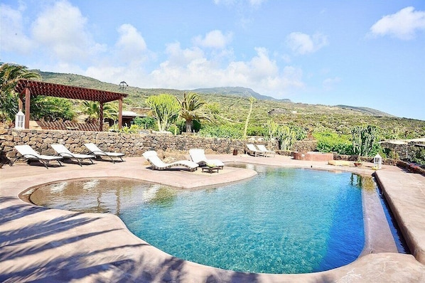 Relax by the serene pool surrounded by a low wall and lounge chairs, shaded by a pergola.