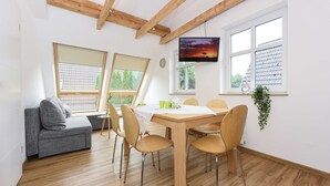 Uma sala de jantar com mesa e cadeiras de madeira, um sofá cinzento e uma TV fixada na parede.