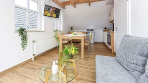 Uma sala moderna com uma mesa de centro de vidro, um sofá cinza, piso de madeira, um espaço de refeições com mesa e cadeiras, uma cozinha com eletrodomésticos e uma grande janela com persianas.