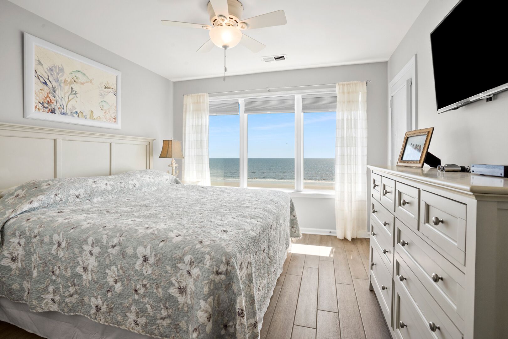 A bedroom with a large bed, a ceiling fan, a dresser, a TV, and a window with a view of the sea.