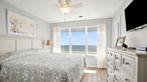 Une chambre avec un grand lit, un ventilateur de plafond, une commode, un téléviseur et une fenêtre donnant sur la mer.