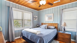 Een slaapkamer met een plafondventilator van hout, een bed met een blauw-witte dekbedovertrek, twee kussens, een nachtkastje met een lamp en een raam met jaloezieën.