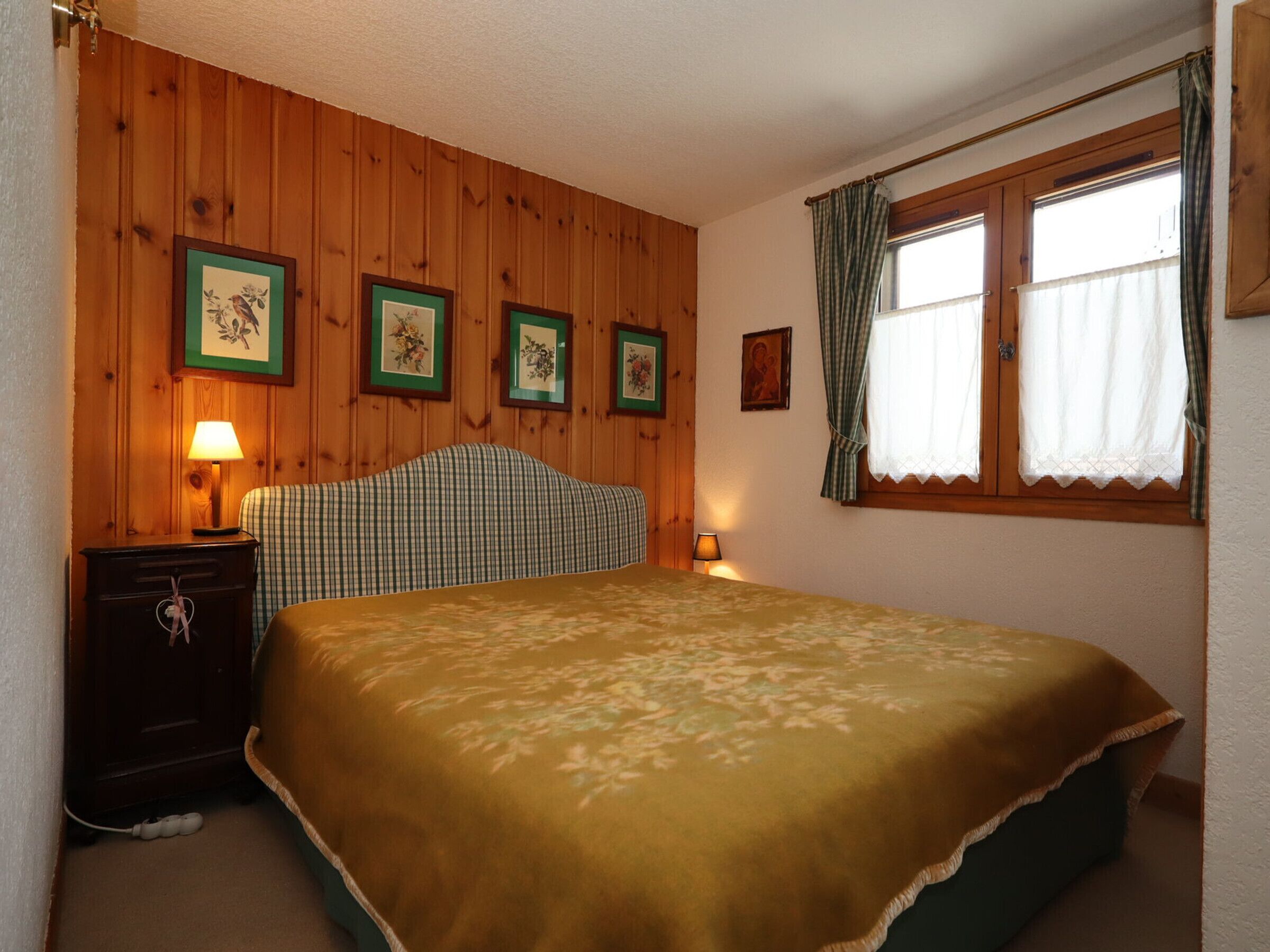 A bedroom with wooden panel walls, a bed with a green checkered headboard, a bedside table with a lamp, and a window with curtains.