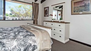 Une chambre à coucher avec un lit, une commode, un miroir et une fenêtre donnant sur des arbres.