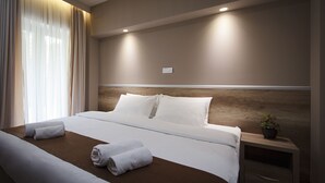 Una habitación de hotel con una cama grande, un cabecero de madera, una mesita de noche con una planta y una ventana con cortinas translúcidas.