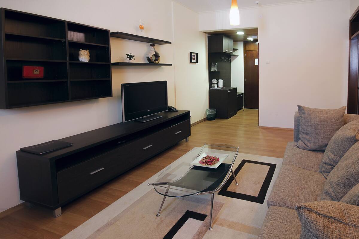 Una sala de estar moderna con un televisor de pantalla plana, mueble de entretenimiento de madera oscura, una mesa de centro de vidrio y un sofá color beige.