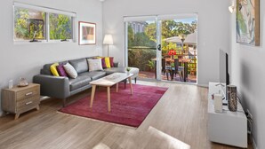 A modern living room with a grey sofa, wooden side table, and a red rug. A sliding glass door leads to an outdoor area with a table and chairs. - Villa 17  Beaches Bush Wineries and more (Dunsborough)