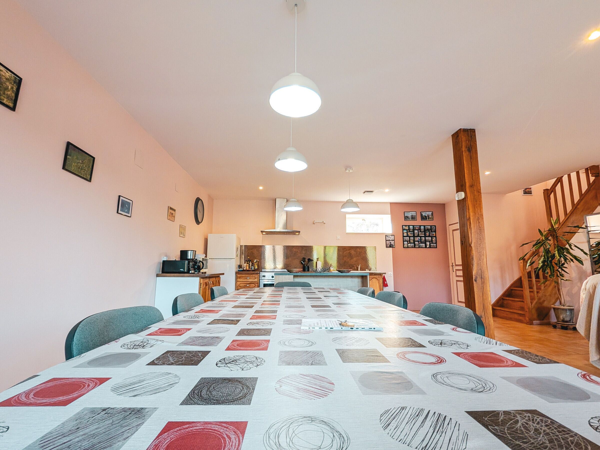 Un comedor espacioso con una mesa larga, sillas, lámparas colgantes, una cocina al fondo y una escalera que sube a otro nivel.