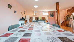 Een ruime eethoek met een lange tafel, stoelen, hanglampen, een keuken op de achtergrond en een trap naar een ander niveau.
