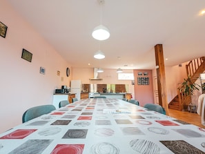 Ein geräumiger Essbereich mit einem langen Tisch, Stühlen, Pendelleuchten, einer Küche im Hintergrund und einer Treppe, die zu einer anderen Ebene führt.