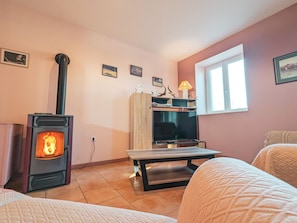 Ein gemütlicher Raum mit Kaminofen, Fernseher auf Holzständer, Fenster und einem Bett mit gemustertem Überwurf.