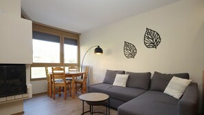 Un salon moderne avec un canapé gris, une petite table ronde et une table à manger avec des chaises. Il y a deux fenêtres qui laissent entrer la lumière naturelle et une lampe fixée au mur.