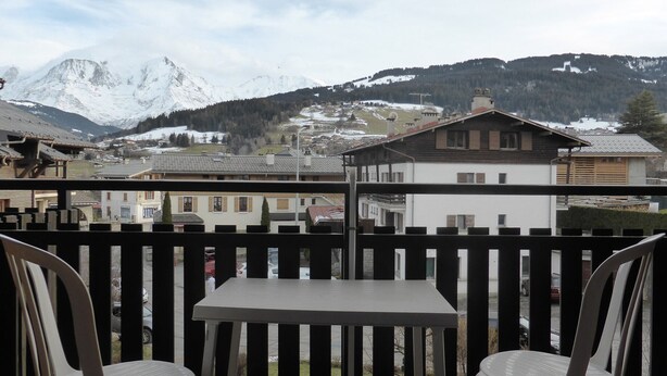 A balcony with two white plastic chairs and a table, overlooking a town with snow-capped mountains in the background.