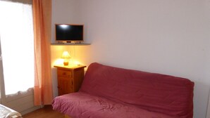 A small room with a wooden floor, a red sofa, a wooden nightstand with a lamp, and a television mounted on the wall.