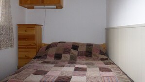 A bedroom with a wooden wardrobe, a single bed with a patchwork quilt, and a plain white wall.