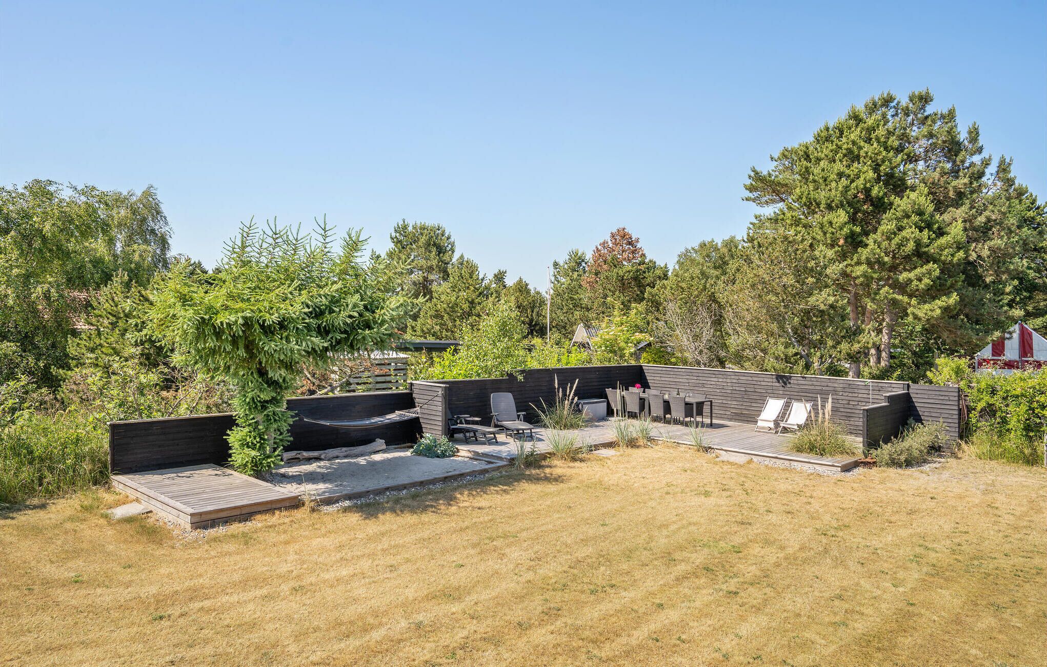 A modern outdoor deck with seating and a barbecue area, surrounded by trees and a grassy lawn.