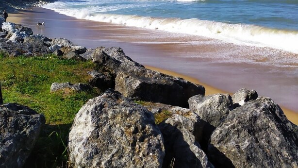 Een strand met grote rotsen op de voorgrond, een zandstrand en golven die op het strand breken.