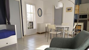 Een woonkamer met een bed, een eettafel, stoelen, een bank, een lamp en een keuken met kasten en apparatuur.