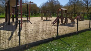 Een speeltuin met diverse speeltoestellen, waaronder glijbanen en klimrekken, omringd door een hek en bomen.