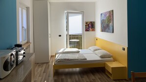 Habitación compacta con cama, lavadora, una mesita y una puerta que da a un balcón.