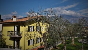 Un edificio amarillo con balcón, rodeado de árboles y montañas al fondo.