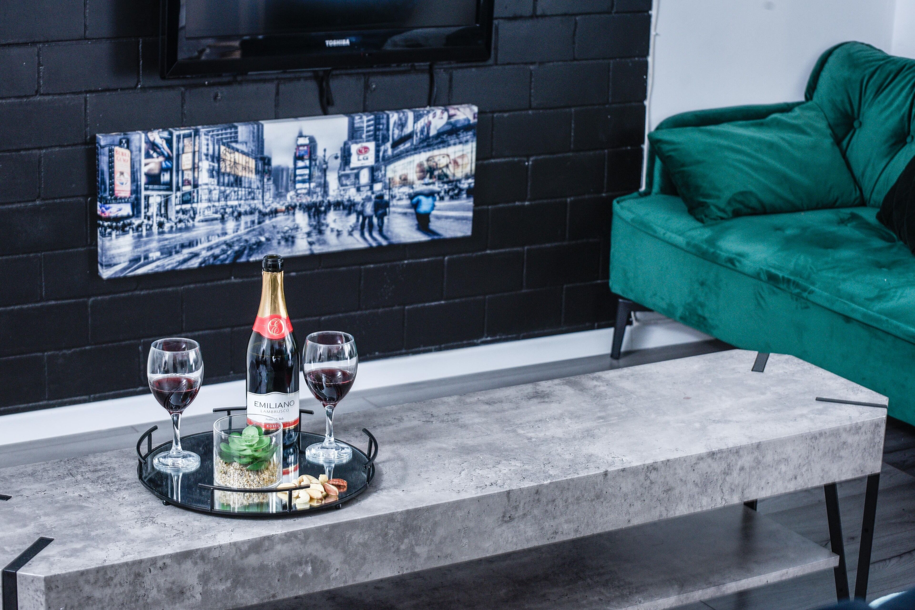 A modern living room with a black brick wall, a green sofa, a mounted flat-screen TV, and a cityscape painting.
