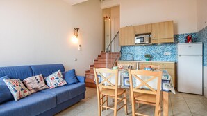 A compact living space with a blue sofa, wooden dining table, and chairs, a kitchen with a microwave and refrigerator, and a staircase leading to an upper level.
