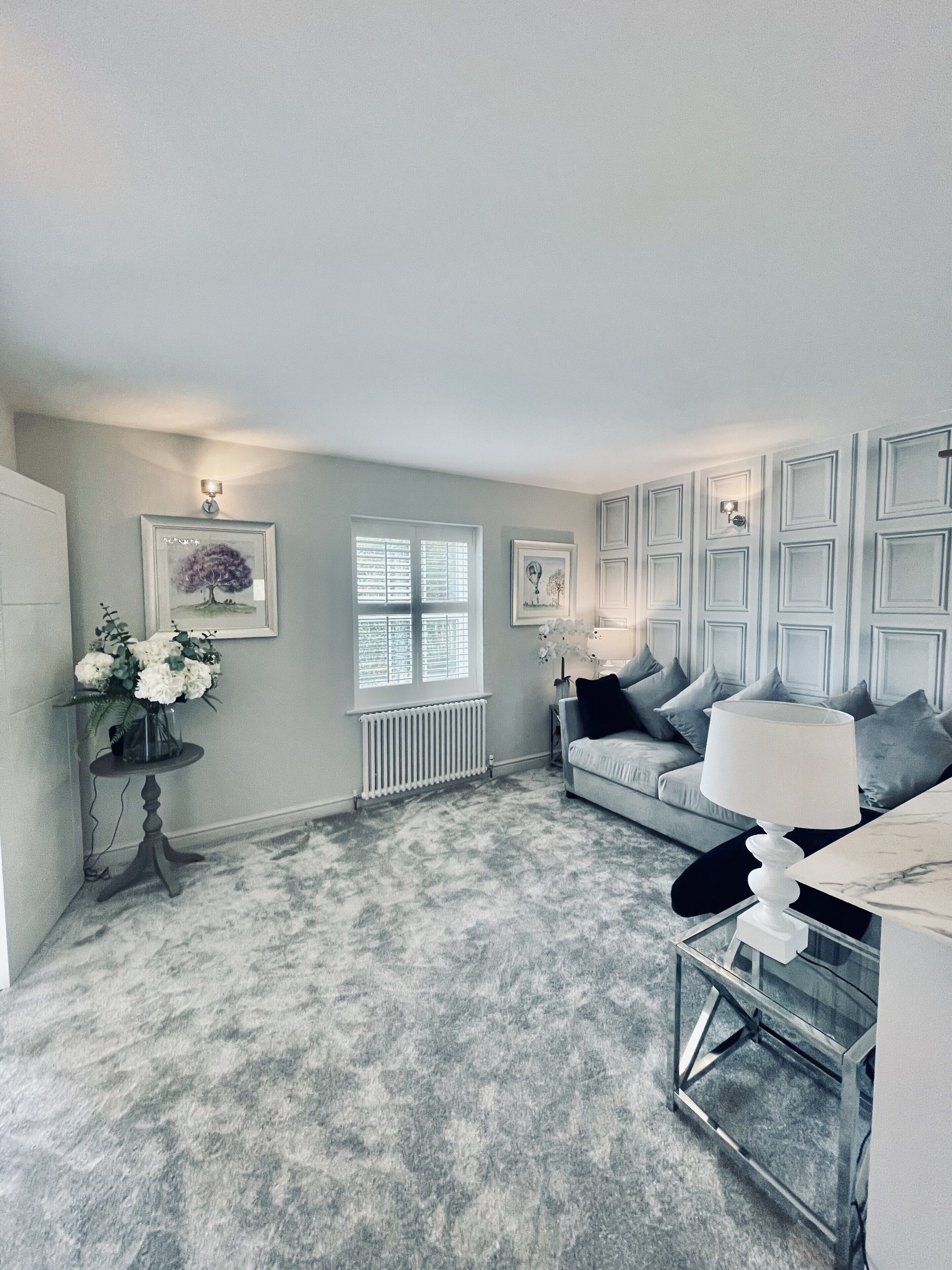 A living room with a grey carpet, a grey sofa with cushions, a coffee table, a side table with a vase of flowers, and framed pictures on the wall.