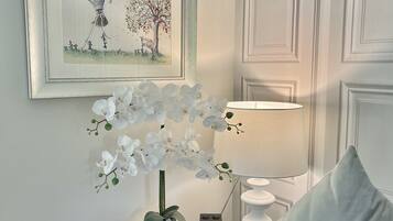 Une chambre avec un lit blanc, un tableau encadré représentant une montgolfière, des orchidées blanches, une table de chevet avec une lampe et une horloge numérique.