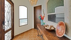 Un pasillo con una puerta de madera, un ventanal grande, una lámpara de araña decorativa y un banco de madera con un jarrón de flores.
